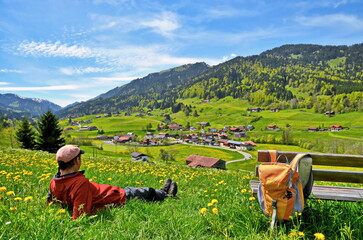 Person, Mann beim Wandern macht Rast in Blumenwiese mit Aussicht auf Berge und Dorf - obrazy, fototapety, plakaty
