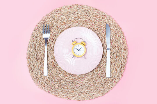Empty plate with alarm clock, fork and knife on pink background, intermittent fasting concept.
