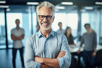 A happy professional man in office with eyeglasses, folded arms and confident expression as other workers hold a meeting in background. - obrazy, fototapety, plakaty