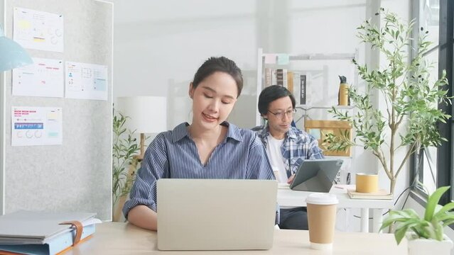 Beautiful young female Asian worker is happy working with laptop, typing, smiling at desk, thinking of ideas, and concentrating creative marketing jobs in white casual office and startup SME business.