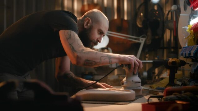 Professional male craftsman grinds wooden guitar body on table using grinding machine. Woodworking specialist, carpenter makes musical instrument in modern workshop. Handicraft and small business.