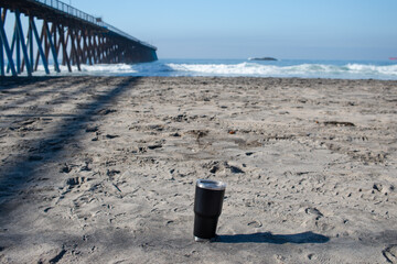 Black metal thermos cup embedded in the sand on the beach. reusable coffee cup