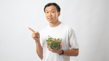 Portrait of an Indonesian Asian man, wearing a white T-shirt, joyfully posing while eating salad from a large transparent bowl, isolated against a white background.