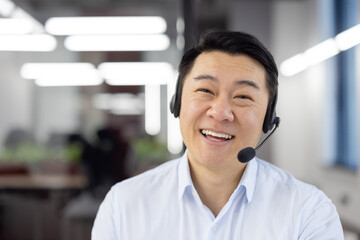 Webcam view, Asian support worker in shirt with headset smiling and looking at camera, businessman working inside office, consulting clients remotely, remote meeting with colleagues partners. - Powered by Adobe