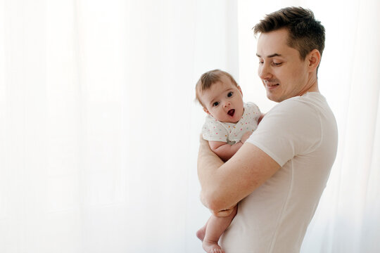 Side view of happy young father in casual clothes with dark hair holding cute baby while standing near white curtain spending leisure time in light room at home