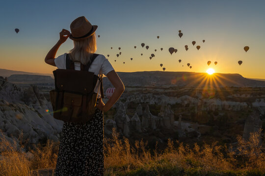 Young female tourist in a hat with a backpack admires the amazing scenery of hot air balloon flights over the valley of love at the moment of a beautiful sunrise, Cappadocia, Turkey.