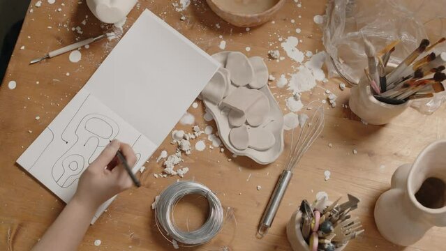Top view of young female sculptor drawing abstraction in sketchbook sitting at messy wooden desk with clay items and stains on it in creative workshop