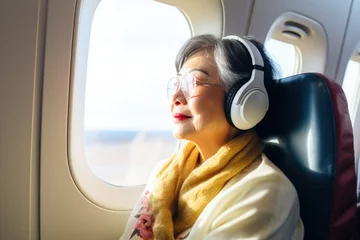 Photo sur Plexiglas Ancien avion An old Asian woman wearing headphones flies on an airplane, sits near the window and listens to music.