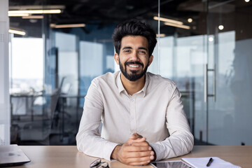 Portrait of a young Indian man in a shirt sitting smiling at a desk in the office and looking confidently at the camera - obrazy, fototapety, plakaty