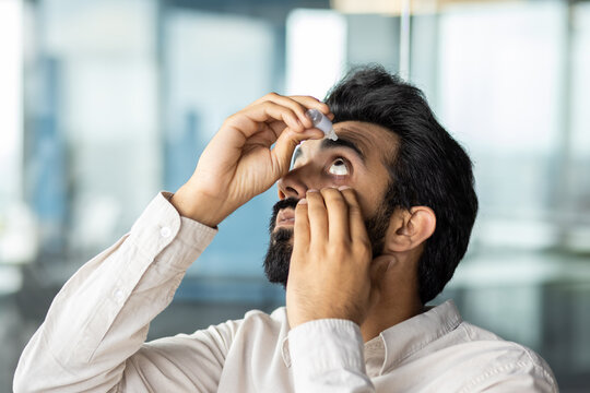 Close-up photo of an Indian young business man working in the office, applying medical drops to his eyes from fatigue and pain
