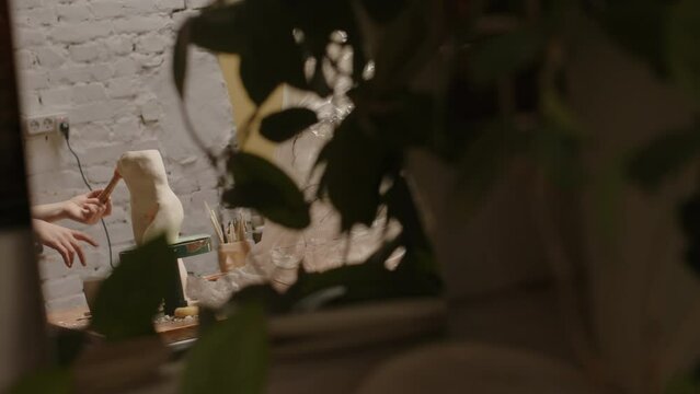 Through potted houseplant shot of young concentrated Caucasian female pottery worker cleaning ceramic female body vase on rotating stand with brush and finishing work
