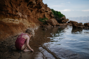 Small girl in swimsuit playing at beach, crouching, seraching for shells in the sand.