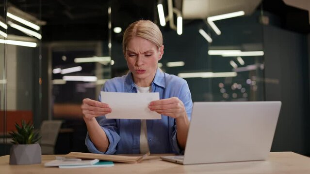 Woman opening letter envelope and getting disappointed while sitting at workplace with laptop in modern office. Blond female receiving notification from boss and learning that she got fired from work.