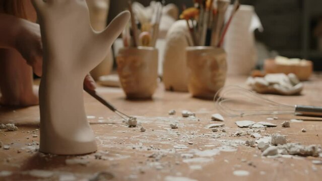 Cropped shot of unrecognizable female pottery worker scraping dry plaster of Paris from wooden work desk after sculpting