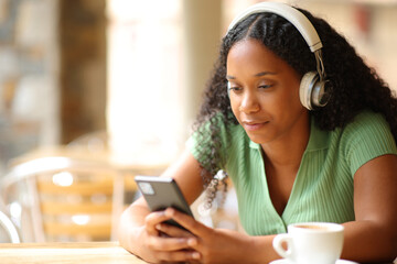 Black woman listening to music using phone and headphone in a bar - Powered by Adobe