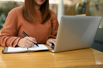 Young female freelancer working with laptop and writing details on notebook