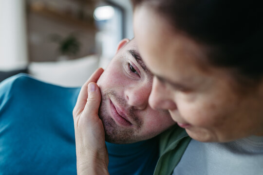 Portrait of young man with Down syndrome with his mother at home, holding, hugging. Concept of love and parenting disabled child.