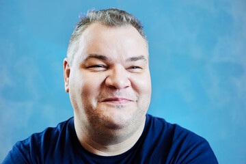 Fototapeta premium Mysterious smile of fat white man in her 40s, face close-up on blue background.