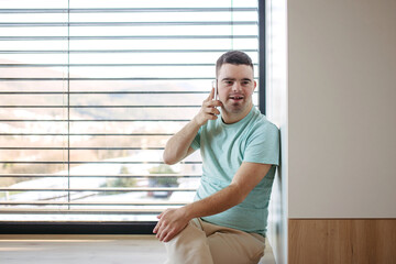 Fototapeta premium Young man with Down syndrome sitting by window, making phonecall with smartphone.