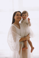 Happy mother in white outfit standing near window and holding her baby daughter in her arms. Isolated on white