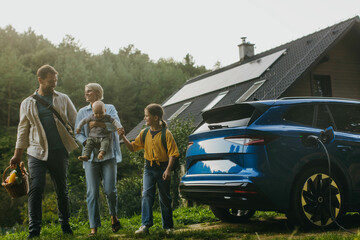 Family with electric car standing in front their house with solar panels on roof. Solar energy and sustainable lifestyle of young family. Concept of green energy and sustainable future for next