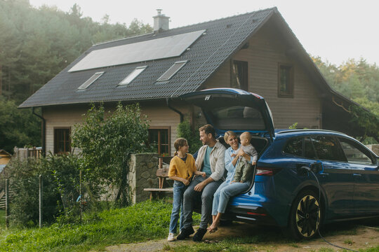 Family with electric car standing in front their house with solar panels on roof. Solar energy and sustainable lifestyle of young family. Concept of green energy and sustainable future for next