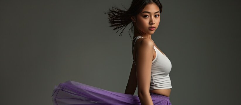 A sensual Asian female model strikes a pose in a photo studio wearing a white tank top and purple skirt against a grey isolated background.