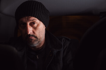 Serious worried man thinking at back seat of the car at night. Adult male wearing black cap and jacket and thinking.
