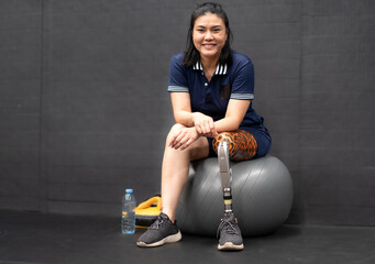 Woman with prosthetic leg sits relaxing on ball in gym. Asian female with foot prosthesis physical...