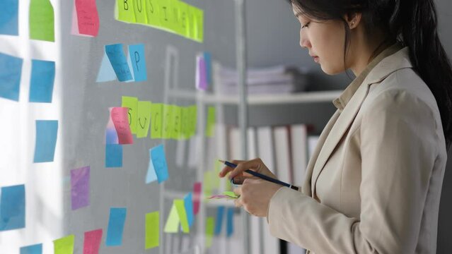 Creative young Asian woman thinking and planning a startup business with paper stickers on a transparent board. Attractive businesswoman analyzing strategy and planning business in office.