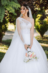 Wedding photo. A brunette bride in a lacy voluminous dress, holding a bouquet and walking in the garden, smiling sincerely. Gorgeous hair and makeup. Fashion and style. Celebration.