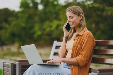 Young woman wear orange shirt casual clothes hold use work on laptop pc computer talk on mobile cell phone sit on bench walk rest relax in spring city park outdoors on nature. Urban lifestyle concept