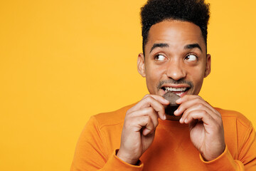 Young man wear orange sweatshirt casual clothes hold eat piece of chocolate look aside isolated on plain yellow background studio portrait. Proper nutrition healthy fast food unhealthy choice concept.
