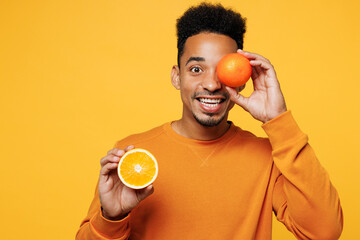 Young smiling man wear sweatshirt casual clothes hold in hand half of orange cover eye isolated on plain yellow background studio portrait. Proper nutrition healthy fast food unhealthy choice concept.