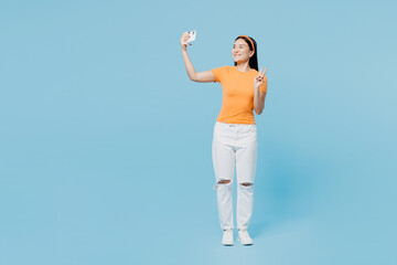 Full body young woman of Asian ethnicity wear orange t-shirt casual clothes do selfie shot on mobile cell phone post photo on social network show v-sign isolated on plain pastel blue cyan background. - obrazy, fototapety, plakaty