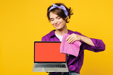 Young IT woman wear purple shirt hold basin with detergent bottles do housework tidy up wiping blank screen laptop pc computer with dust cloth isolated on plain yellow background Housekeeping concept