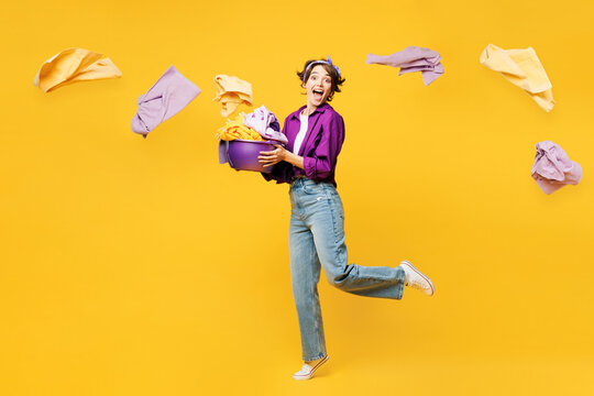 Full body side view young woman wear casual purple shirt do housework tidy up hold basin with laundry near flying clothes look camera isolated on plain yellow background studio. Housekeeping concept.