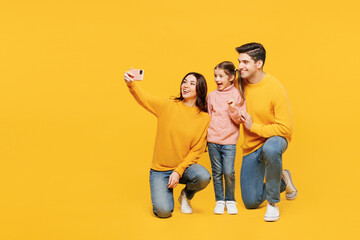 Full body young happy parents mom dad with child kid girl 7-8 years old wear pink sweater casual clothes doing selfie shot on mobile cell phone isolated on plain yellow background. Family day concept.