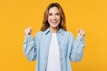 Young happy cool woman she wear blue shirt white t-shirt casual clothes doing winner gesture celebrate clenching fists say yes isolated on plain yellow background studio portrait. Lifestyle concept. - obrazy, fototapety, plakaty