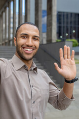 Hispanic smiling young man standing near office building and talking on camera phone, greeting and waving. Vertical photo. © Liubomir