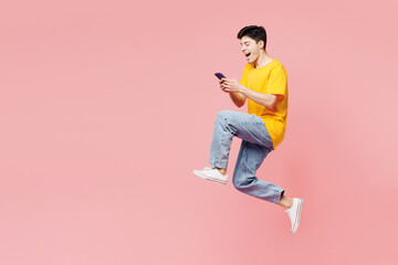 Full body side profile view excited young man wear yellow t-shirt casual clothes jump high hold use mobile cell phone isolated on plain pastel light pink background studio portrait. Lifestyle concept