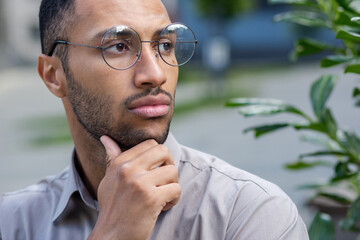 A close-up photo of a young Hispanic man standing outside the city wearing glasses, rubbing his beard with his hand, looking seriously and thoughtfully to the side.