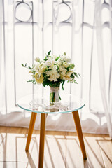 Bridal bouquet stands on a glass table in a room near the white curtain