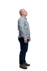 A bald man stands and looks up. A guy in a gray shirt and blue trousers. Full height. Side view. Isolated on a white background. Vertkial.