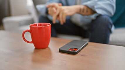 Intimate glance at a man's hands, sitting on a sofa, engrossed with his cellphone, in the cozy home interior. nearby, on the table, sits a cup of his favored beverage.