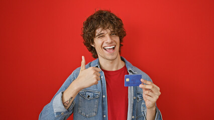 A cheerful young man with a beard giving thumbs up and holding a credit card against a red wall.