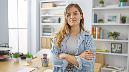 Confident young blonde woman exudes joy & success at her workplace. with a positive smile, she stands, arms crossed, a happy, relaxed & elegant boss in her office room.