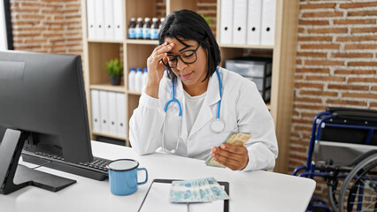 Worried hispanic woman doctor in clinic holding brazilian real currency, with wheelchair in...