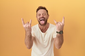 Middle age man with beard standing over yellow background shouting with crazy expression doing rock...