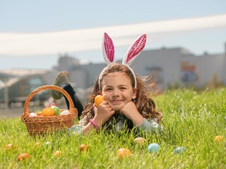 A beautiful little brunette girl in bunny ears is lying on the green grass with a basket full of...